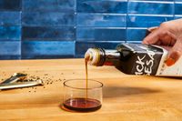 Pouring sesame oil into a small glass container, measuring spoons and sesame seeds nearby on a wooden surface