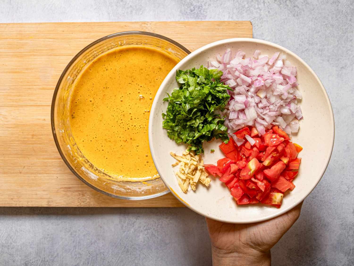 A hand holding a plate with chopped onion tomato coriander and ginger above a bowl of yellow liquid batter