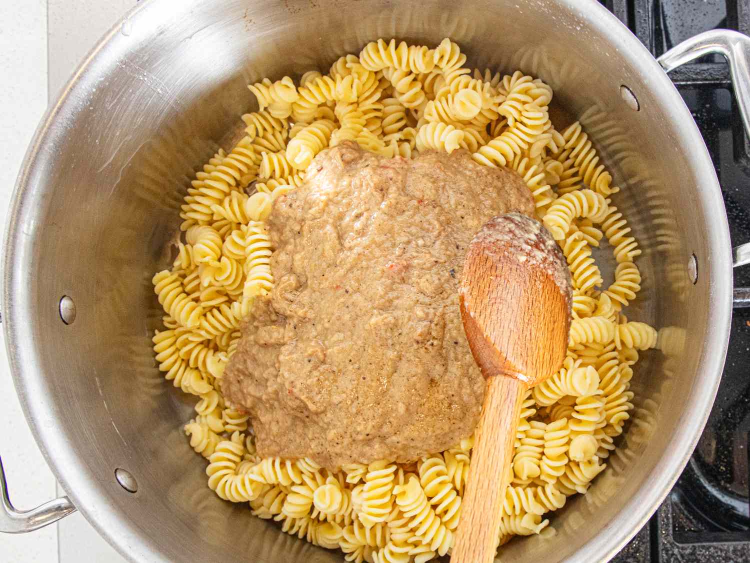 A pot with cooked pasta and baba ghanoush being stirred with a wooden spoon
