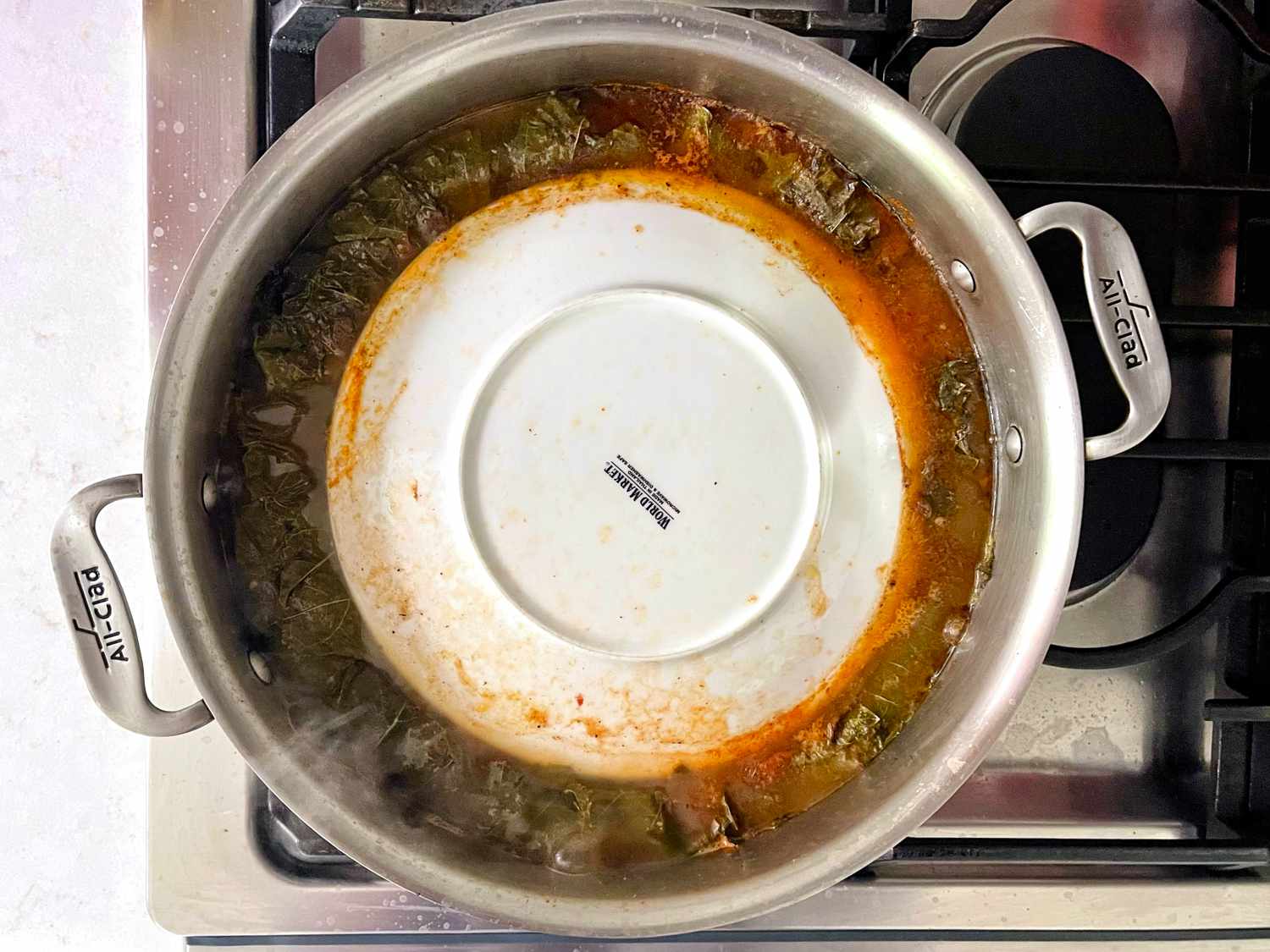 Overhead view of broth bubbling in pot with grape leaves and inverted plate weighing it down
