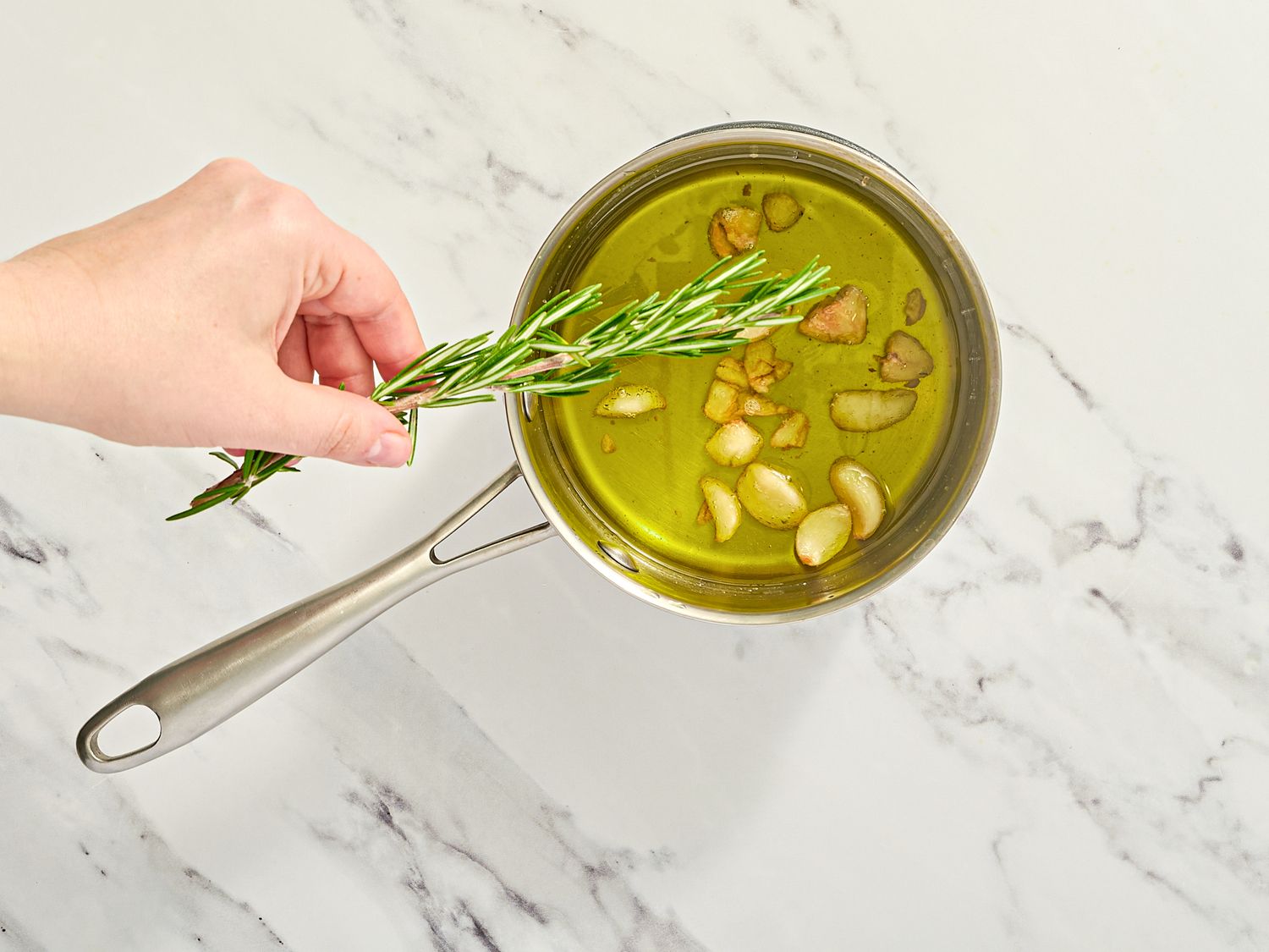A hand adding a sprig of rosemary to a saucepan of oil with garlic cloves on a marble countertop