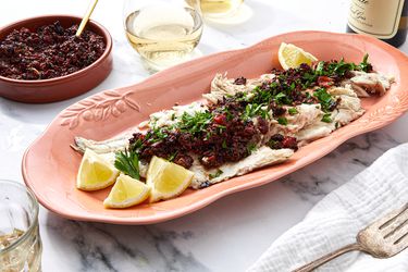 A dish of grilled fish topped with olive and tomato compote served on a table with lemon wedges and parsley garnish