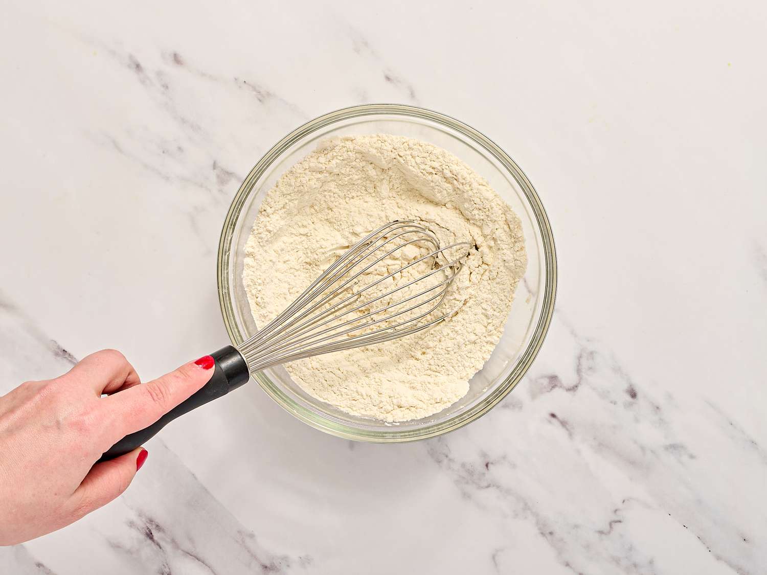 A whisk is being used to mix dry ingredients in a clear bowl on a marble countertop