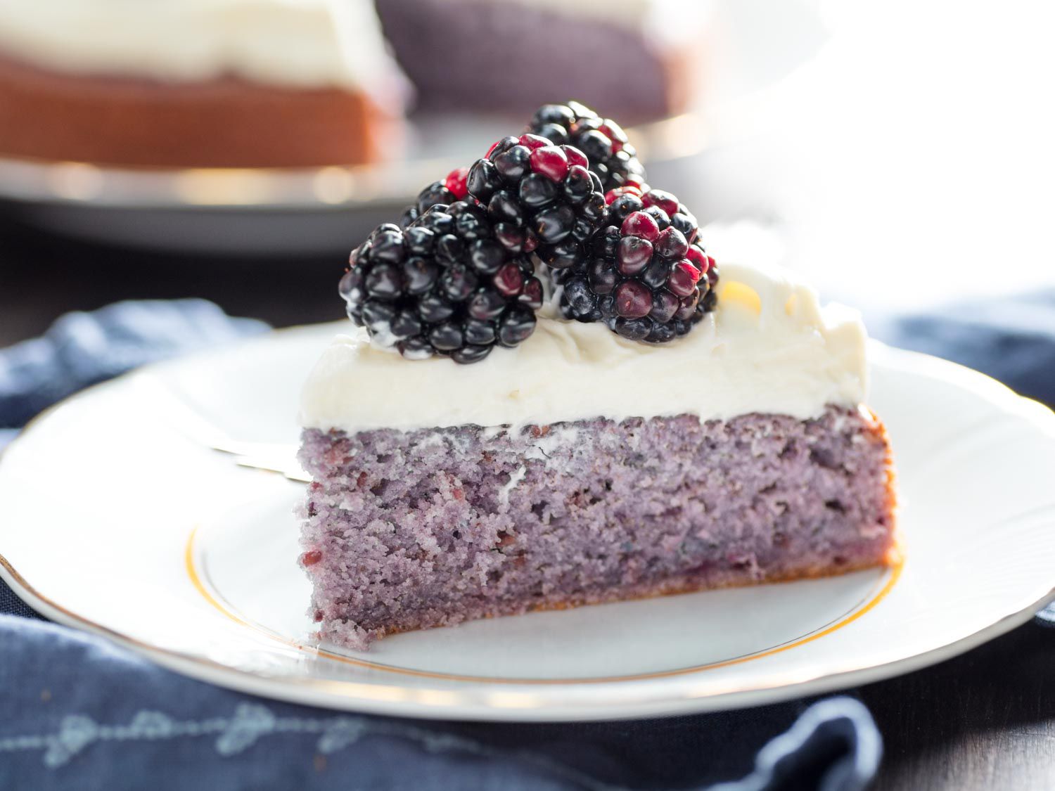 A slice of purplish blackberry cake on a plate, topped with cream cheese frosting and fresh whole blackberries