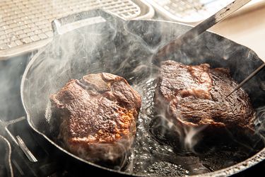 Two steaks cooking in a cast iron skillet with steam rising up