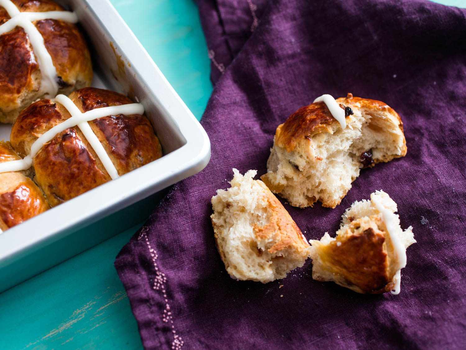 A hot cross bun, torn into pieces, on a purple cloth, next to a dish of other hot cross buns.