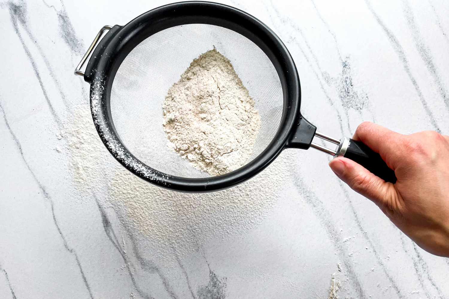 Hand sifting flour through the KitchenAid Classic Strainer on a marble surface