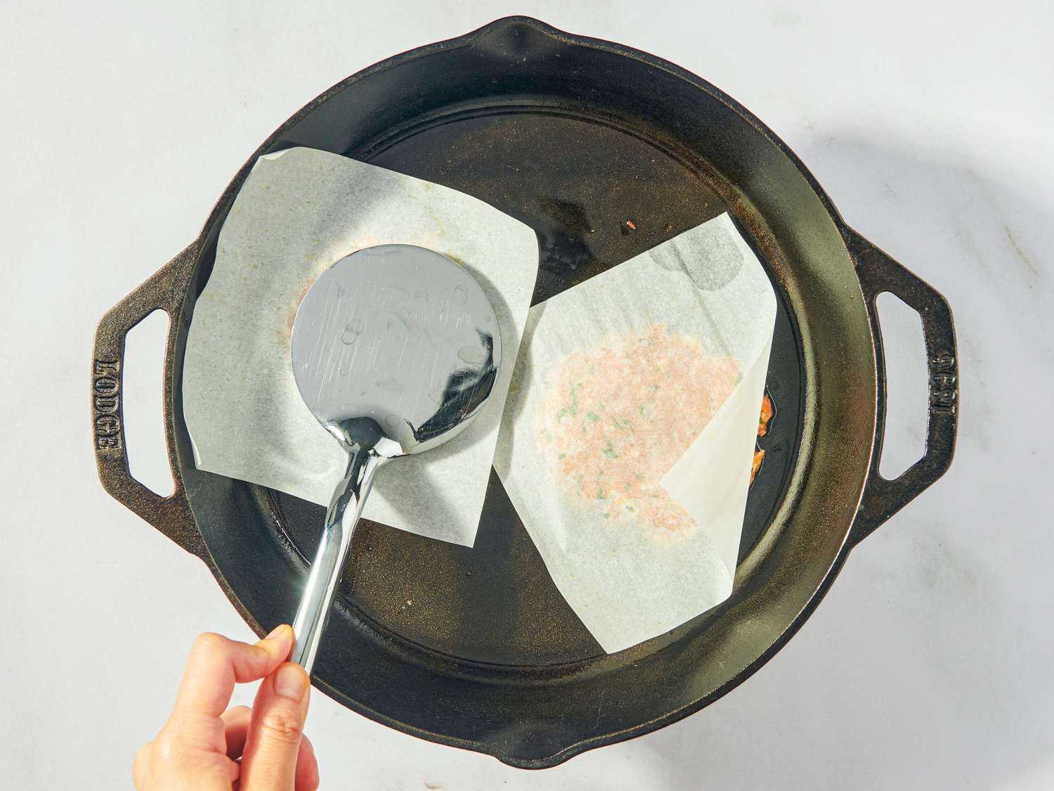 A cast iron pan with parchment paper pieces one being pressed by a hand using a flat spatula