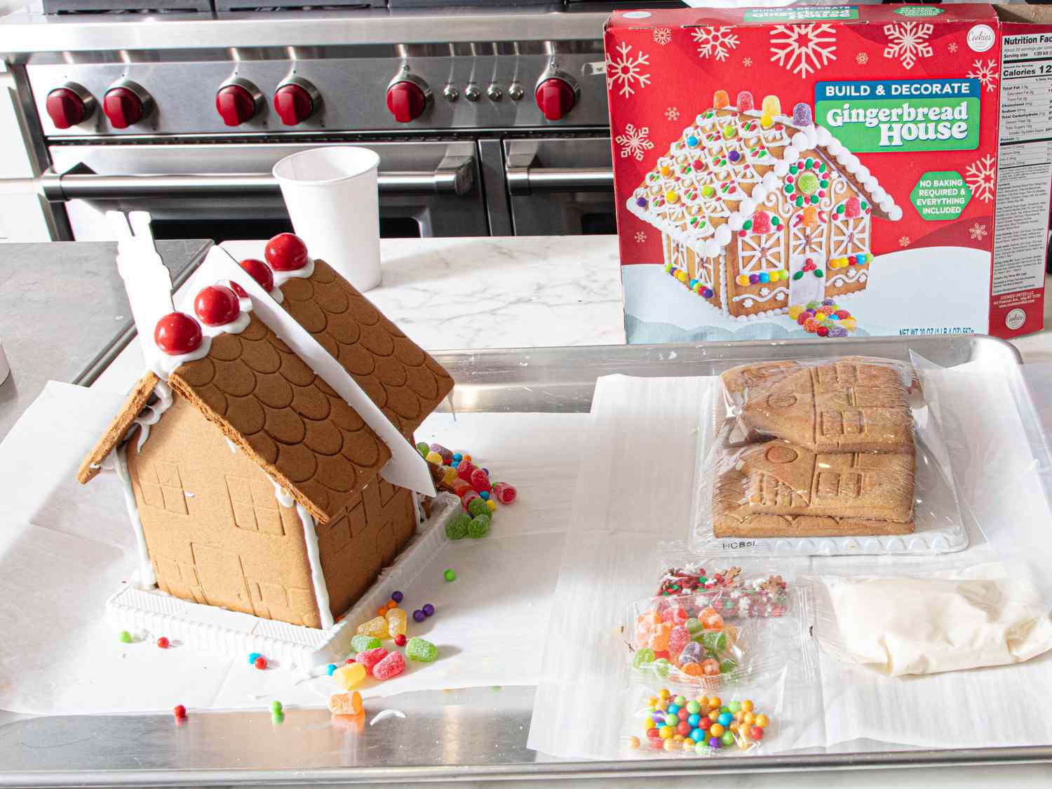 A partially decorated gingerbread house with candies and icing on a counter next to a gingerbread house kit and packaging