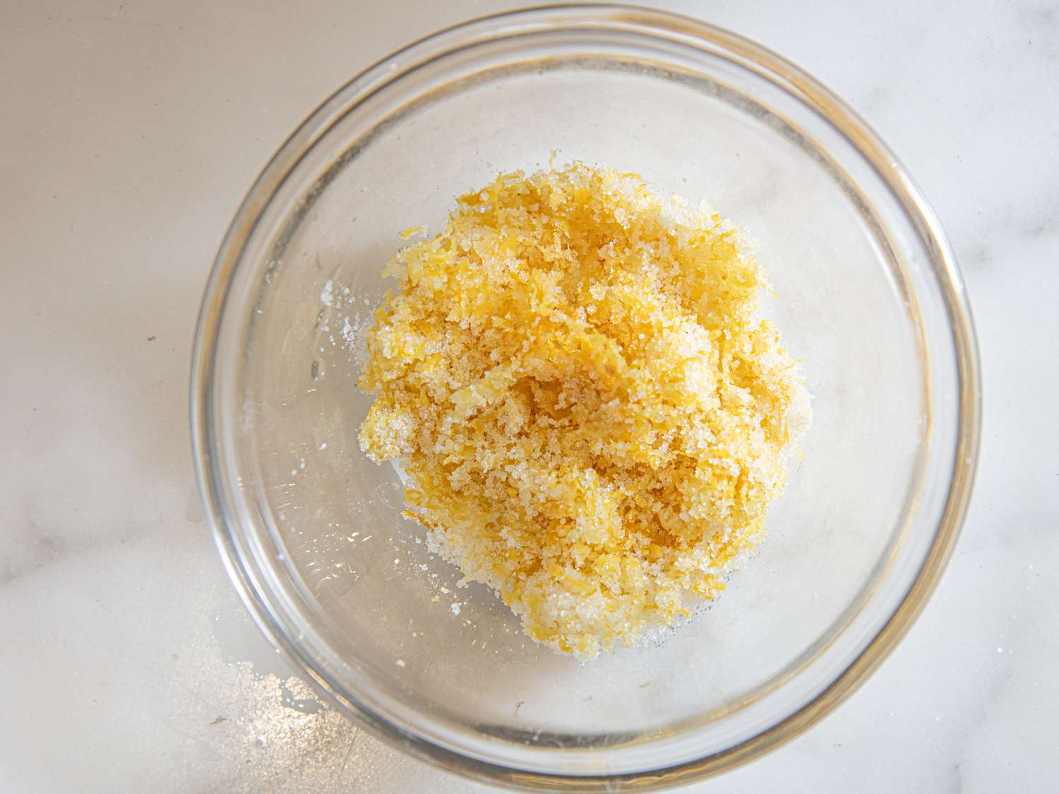 A glass bowl containing grated lemon zest mixed with sugar, prepared for use in a lemon ricotta pancake recipe