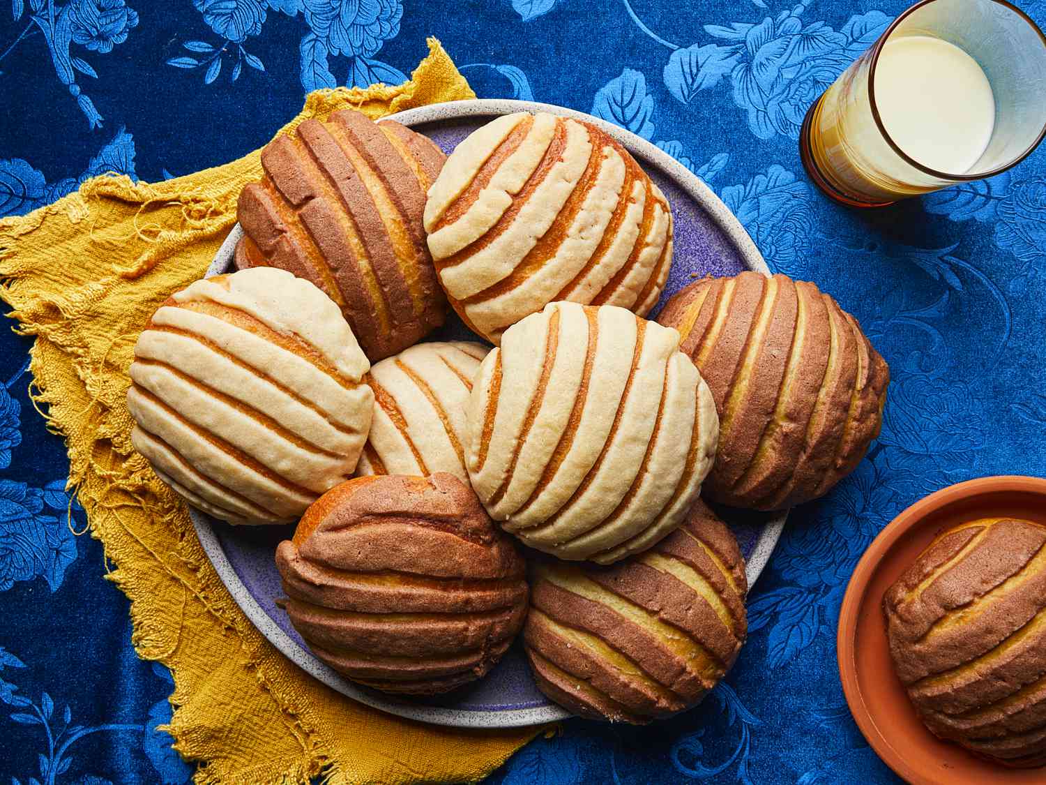 Overhead view of conchas on a plate with a glass of milk. Tablecloth is a velvety blue with a floral pattern and plate is resting on a bright yellow napking