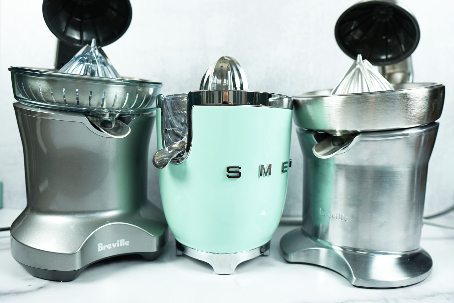 three electric citrus juicers on a marble surface with a grey background