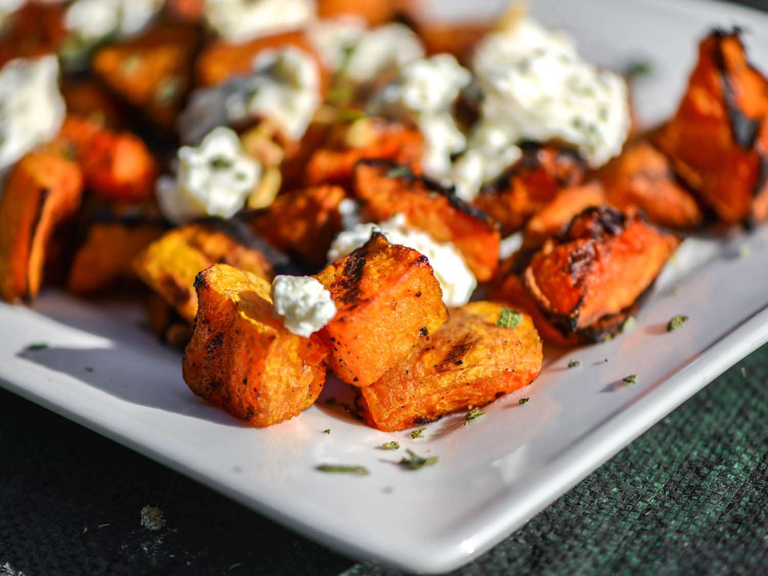 Close-up of grilled butternut squash cubes on a serving plate, garnished with ricotta and sage. 