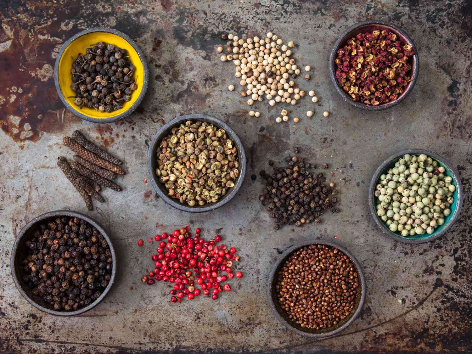 Overhead view of ten peppercorn varieties arranged in bowls or tidy mounds on a seasoned, worn baking sheet.