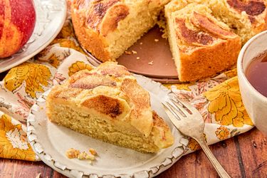 A slice of apple cinnamon cake on a plate part of the cake and an apple shown on a decorative table setting