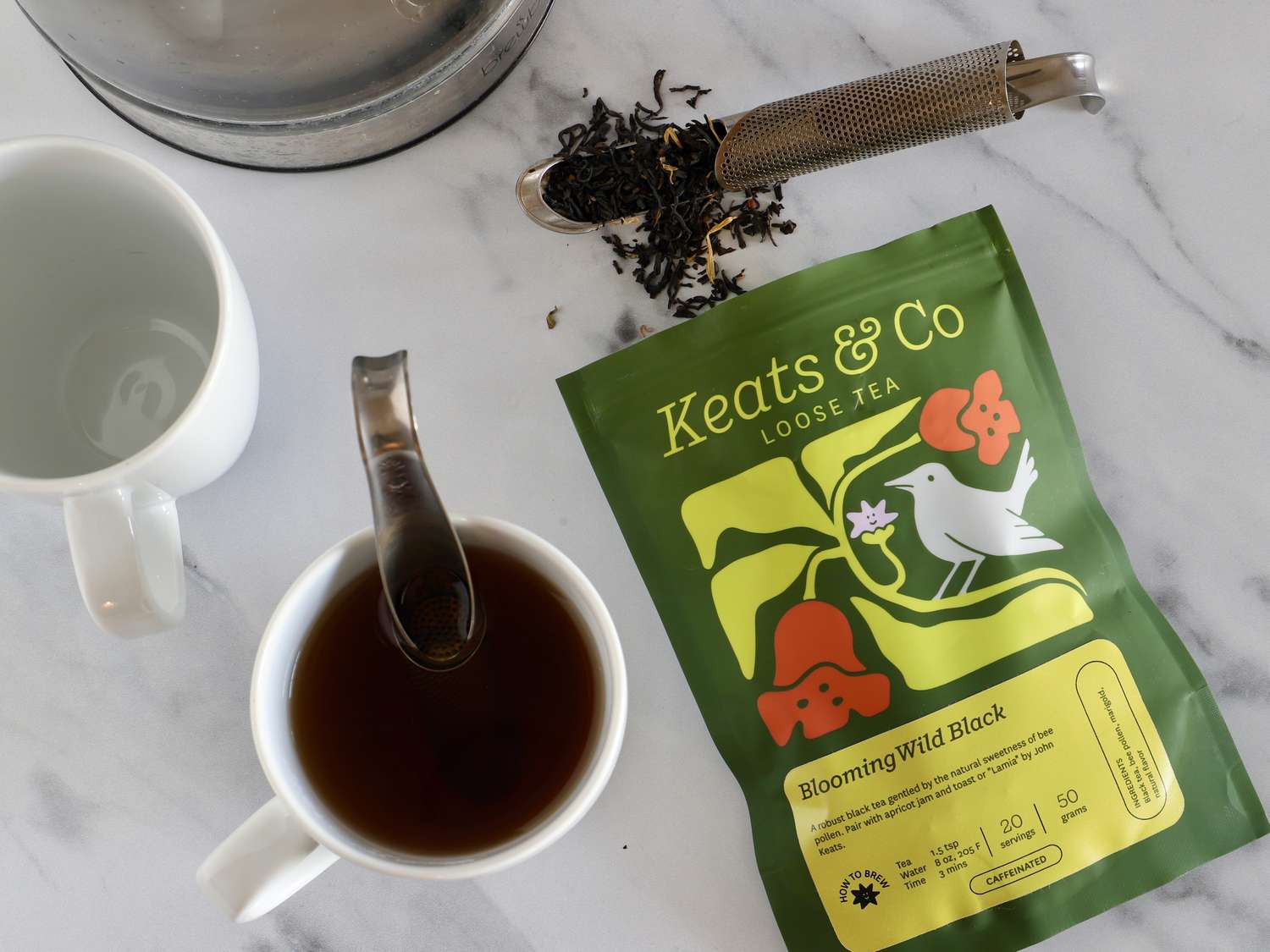 A package of Keats Co blooming wild black loose tea with a brewed cup of tea and a steeped infuser nearby on a marble surface