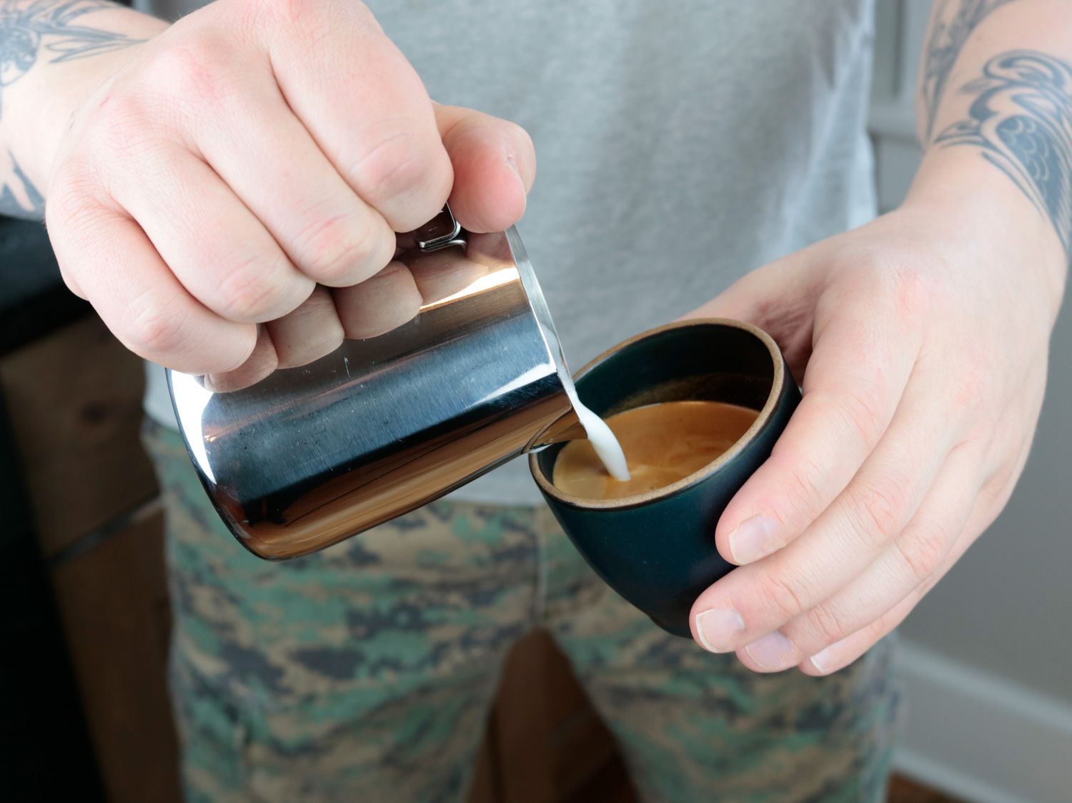 one hand pouring steamed milk from a pitcher into a cup with espresso in it held by the other hand