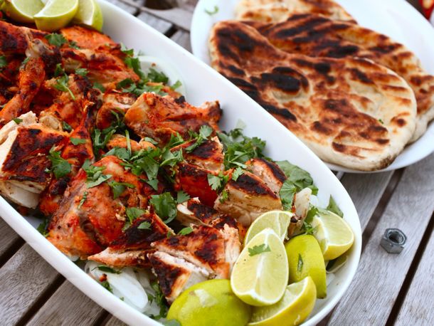 A large platter of tandoor-style Cornish hens cut up into serving pieces and sprinkled with cilantro. Several limes have been cut into wedges and garnish the platter. A plate of deeply browned naan sits next to the Cornish hen.
