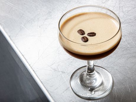 A glass of espresso martini garnished with three coffee beans on a metallic surface