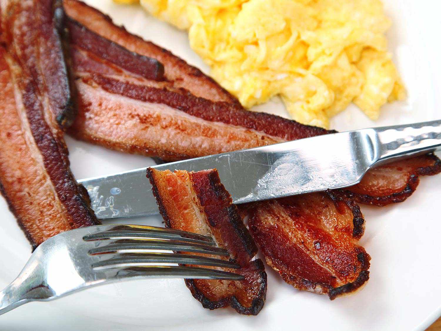 Baked bacon cut up on a plate with scrambled eggs.