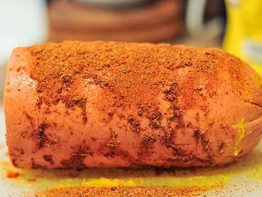The scored, mustard-coated bologna getting sprinkled with a barbecue rub.