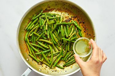 Cooking green beans in a pan with a liquid being poured from a bowl