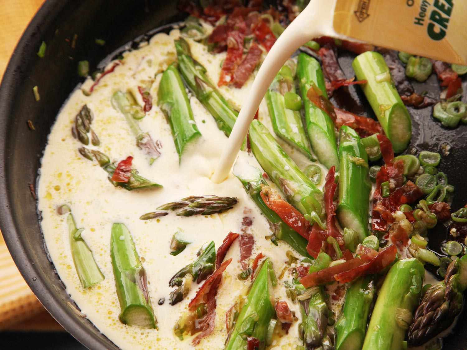 Pouring cream into a skillet with sauted asparagus and prosciutto for a gnocchi dish.