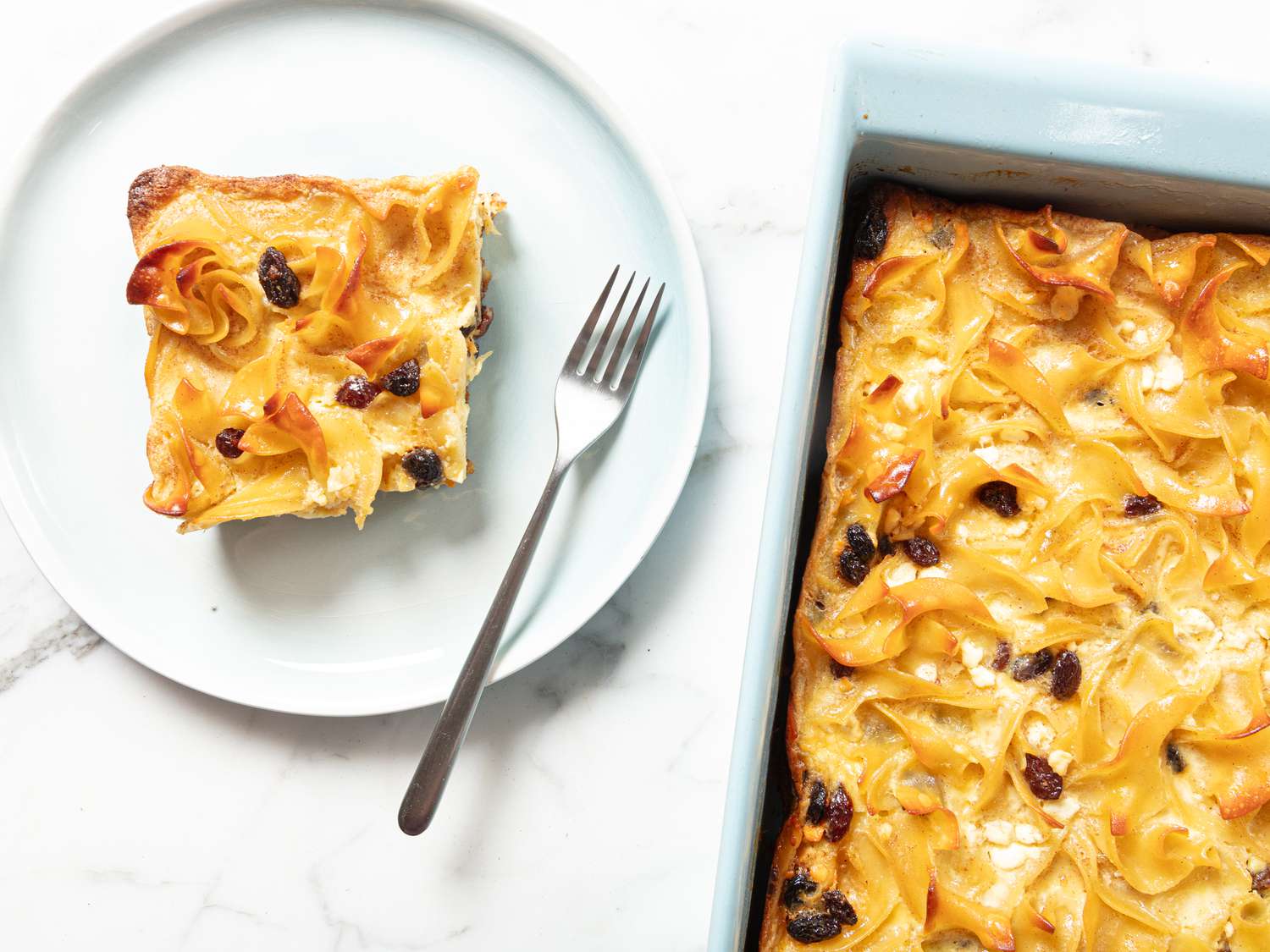 Tray of noodle kugel, Square piece of Noodle Kugel on a light blue plate with a fork on a white marble surface