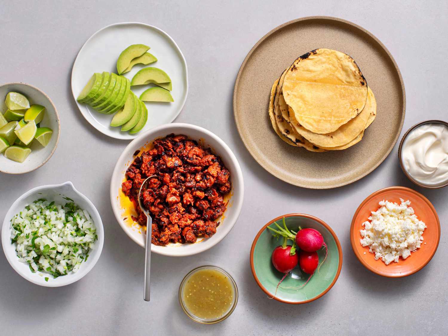 A scene for assembling chorizo tacos: cooked chorizo, charred corn tortillas, sliced avocados, chopped onions and cilantro, lime wedges, radishes, queso fresca, salsa verde, and sour cream