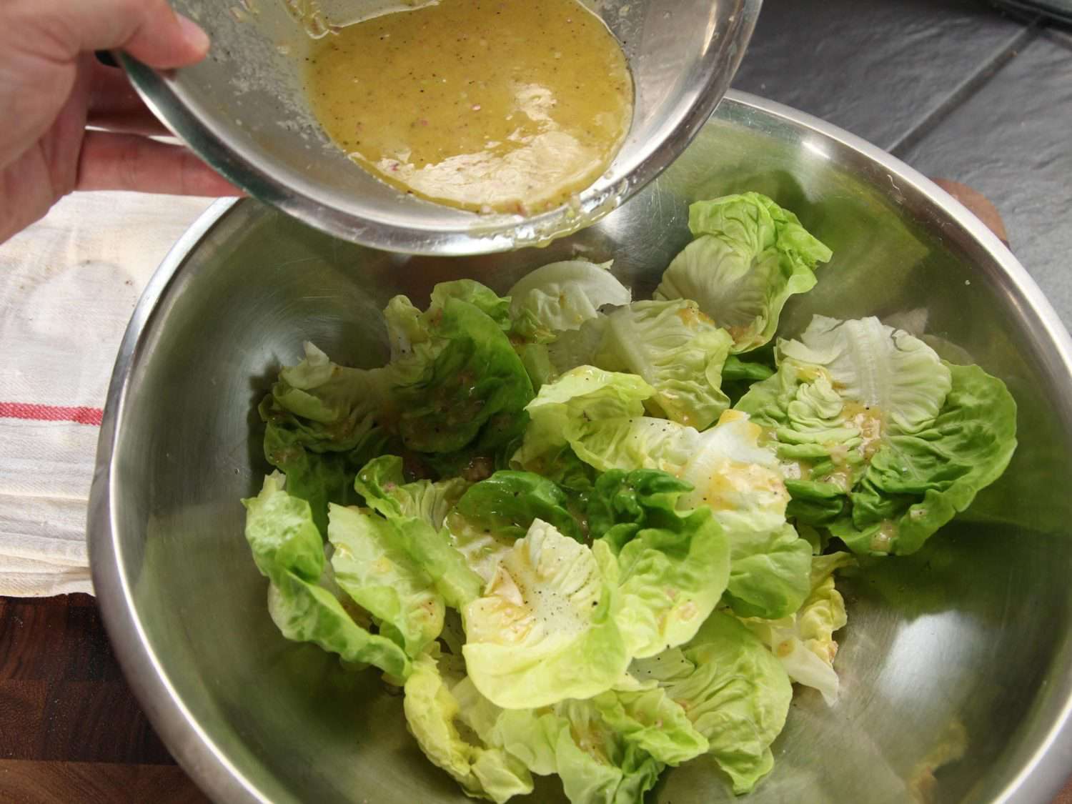 Hand pouring small bowl of vinaigrette over large bowl of salad greens