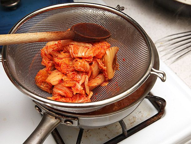 Kimchi being pressed with a wooden spoon on a fine mesh strainer, releasing the juices into the saucepan of dashi.
