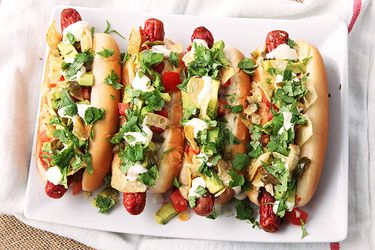 Overhead view of four Bacon-Wrapped Hot Dogs With Avocado, Tomato, Onion, and Potato Chips.