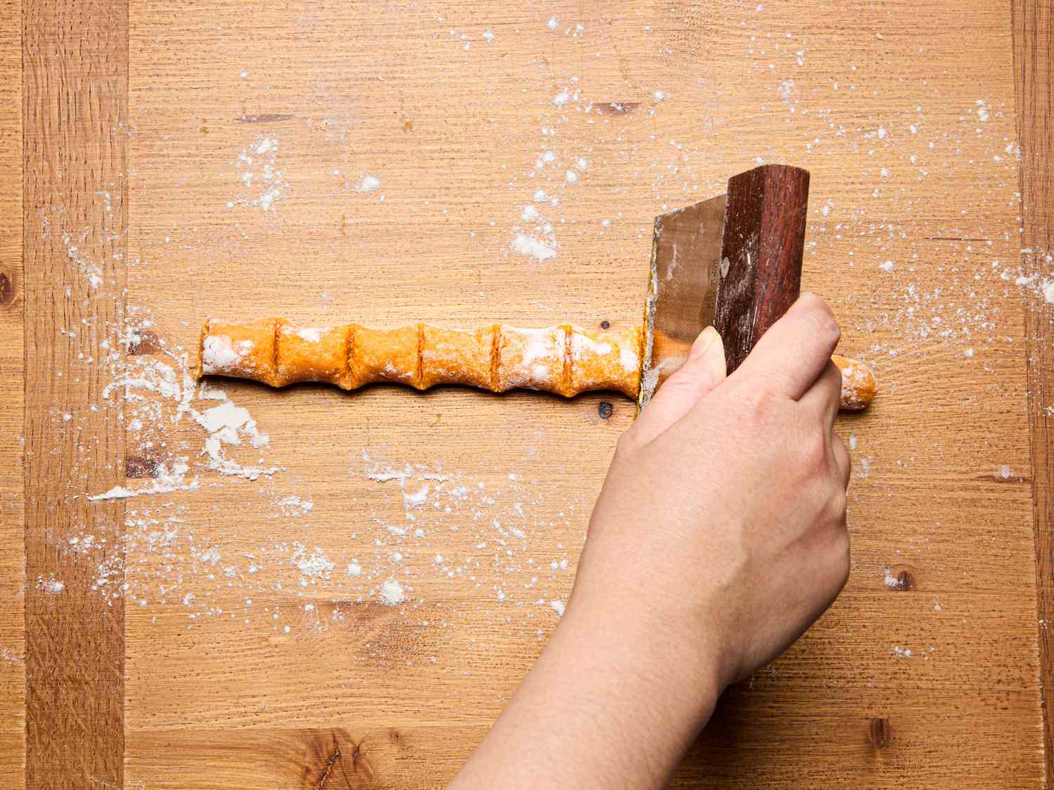 A hand cutting a prepared roll of sweet potato gnocchi dough into individual pieces on a wooden surface