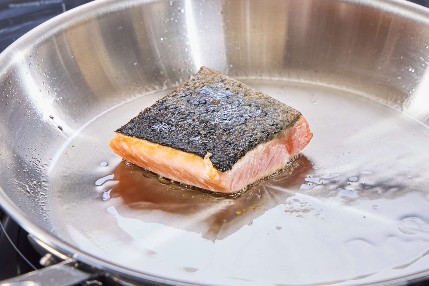 A salmon fillet cooking in a stainless steel skillet.