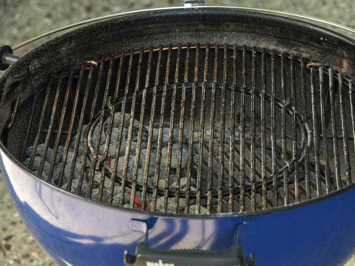 An angled shot of a blue charcoal kettle grill, showing that half of the interior of the grill is layered with hot, ash-covered coals.