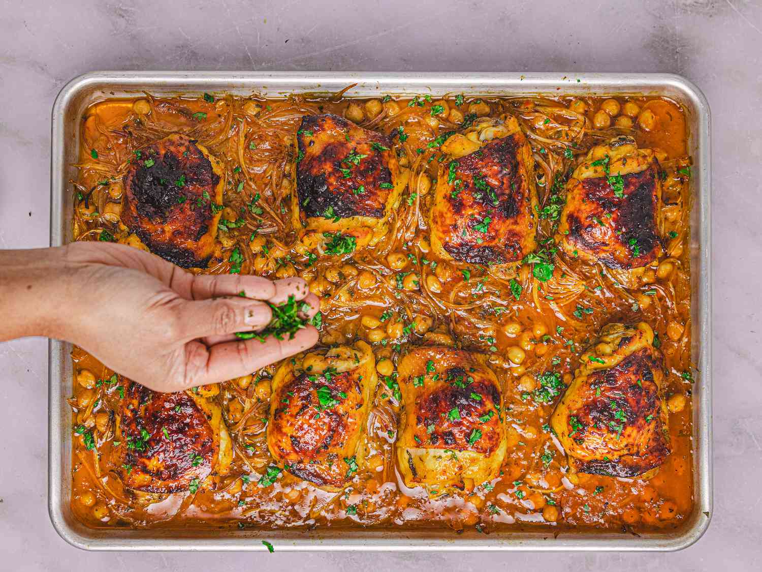 Sheet pan with cooked chicken thighs, chickpeas, and herbs, a hand scattering greens over the dish