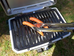 a person using a pair of grill tongs to flip a hot dog on a portable gas grill