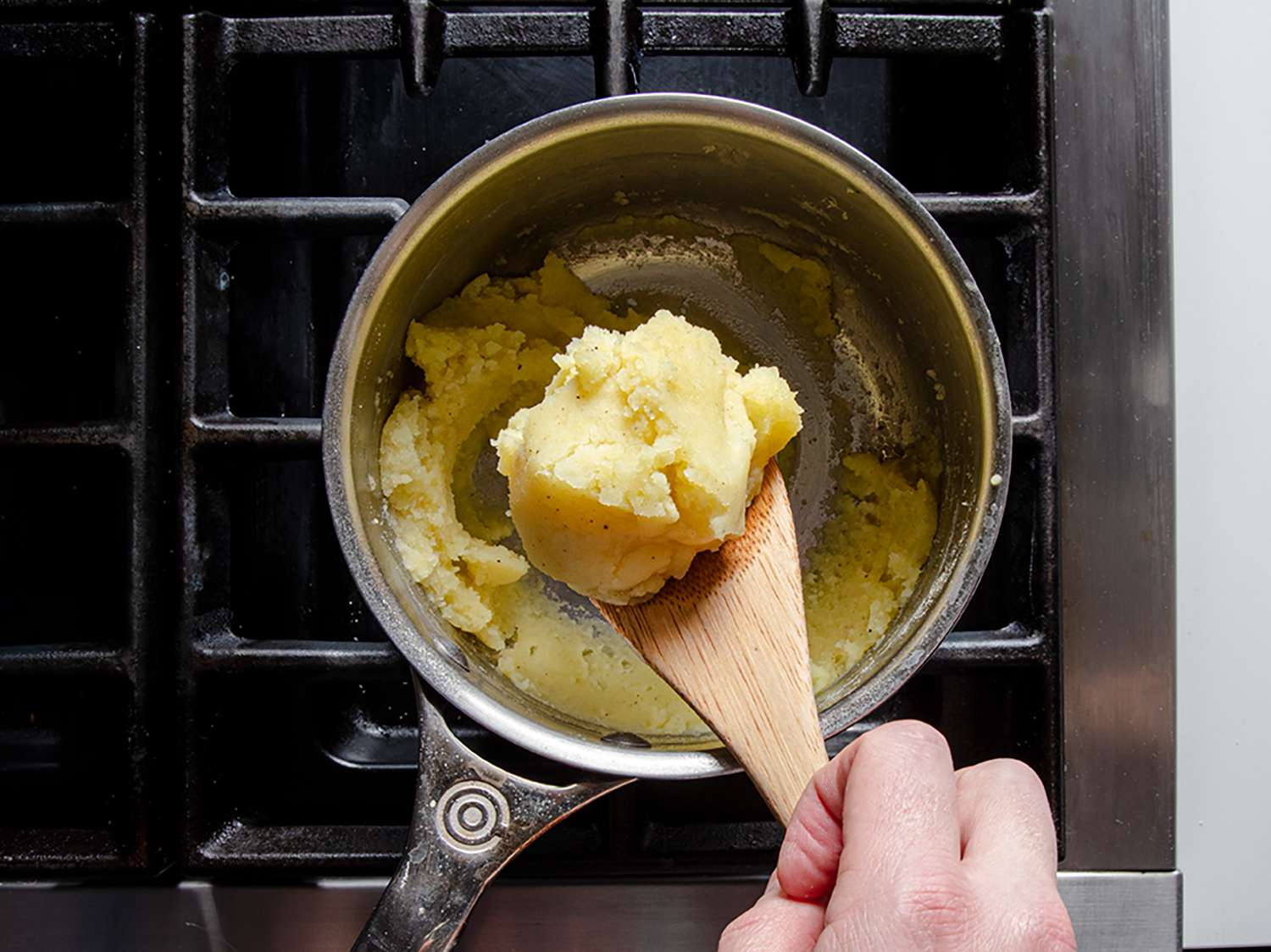 Mashed potatoes being held above a stainless steel pan by a wooden spoon.