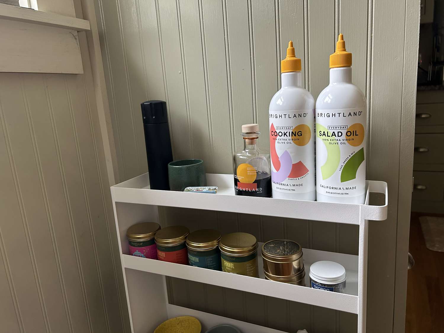 A white rolling kitchen storage rack with olive oil, spices, mugs, napkins, and boxes of pasta on its shelves
