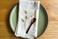 A ceramic plate with a cloth napkin and wooden spoon placed on top, featuring a leaf and flower pattern on the napkin