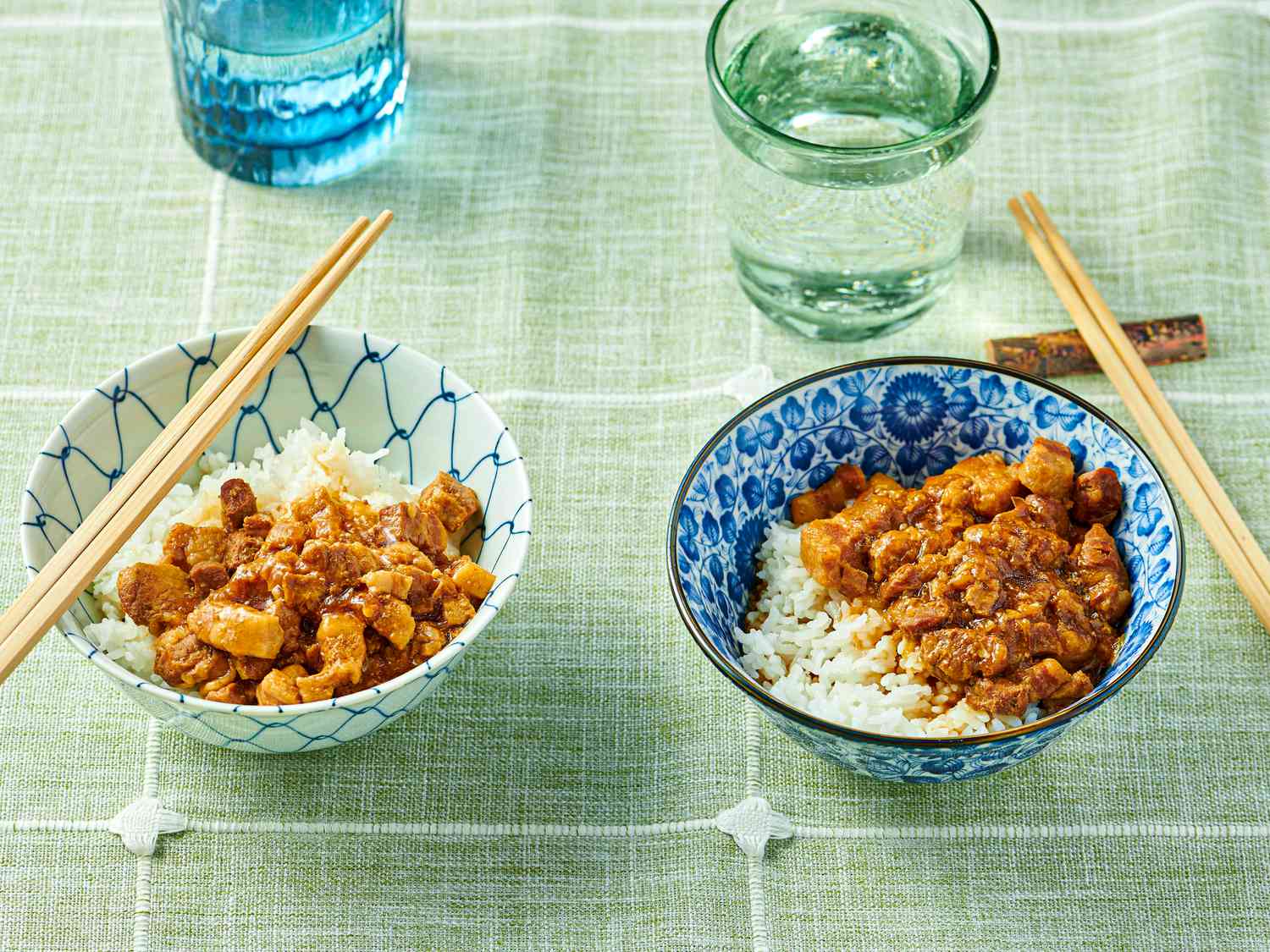 2 bowls of minced pork on white rice in a blue bowl and a white bowl, with chop sticks and glasses of water on a green tablecloth