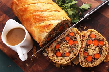 Sliced vegetables Wellington roast resting on cutting board with two slices cut, with gravy pitcher and fresh parsley alongside.