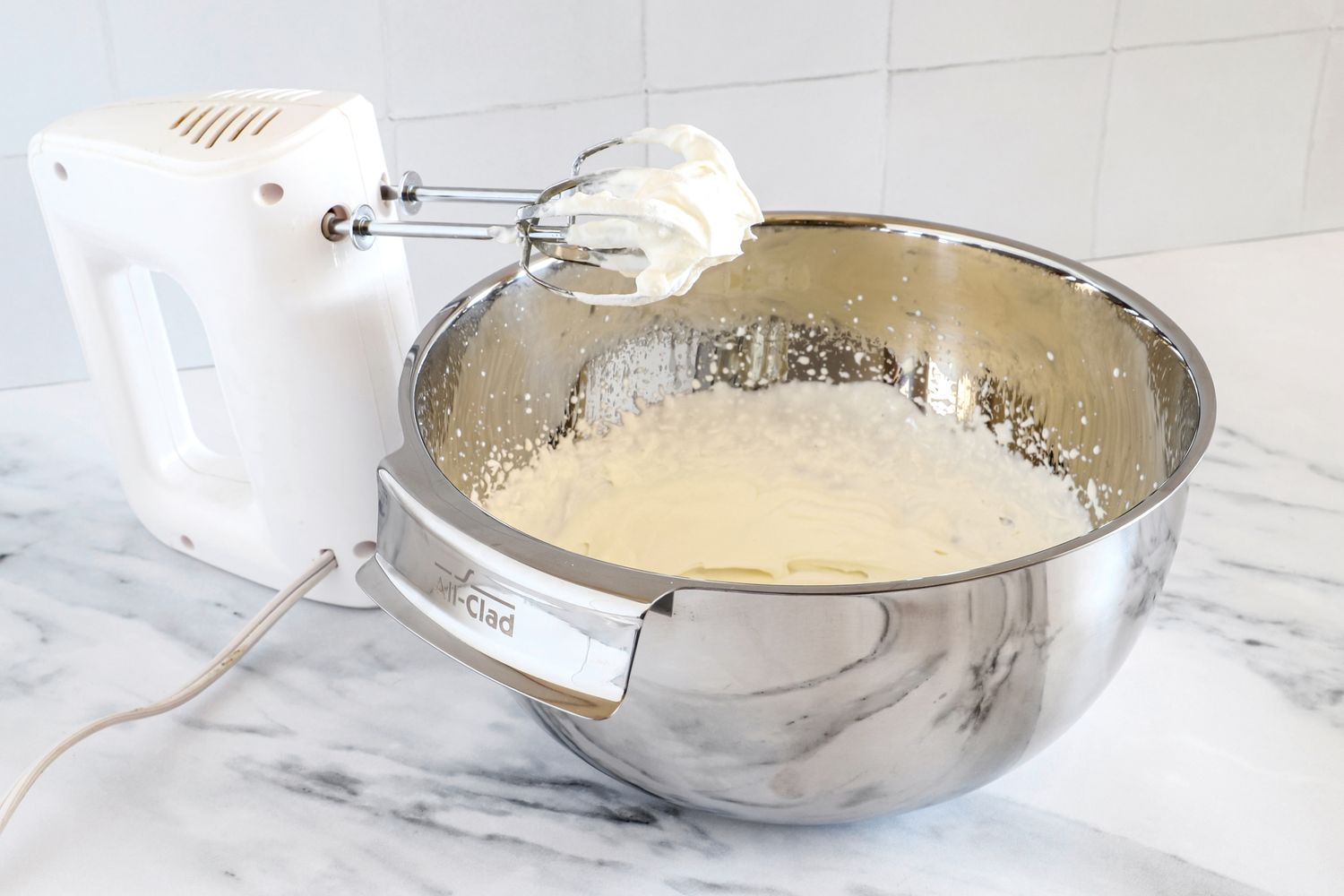 An electric handheld mixer beside an All-Clad mixing bowl containing partially whipped cream