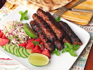 A platter of Pakistani Seekh kebabs with slaw, cucumbers tomatoes, and lime wedges.