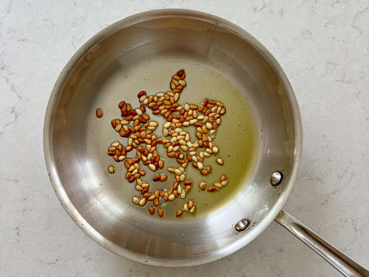 Toasted pine nuts sizzling in a stainless steel frying pan with oil on a lightcolored countertop
