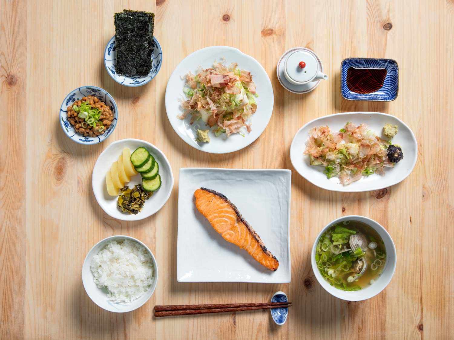 Overhead shot of Japanese breakfast: rice, pickle plate, natto, nori, tofu, soy sauce decanter, soy sauce, eggplant, miso soup, salted salmon on wooden table.