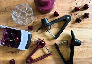 Five cherry pitters on a wooden surface with a few cherries strewn around them