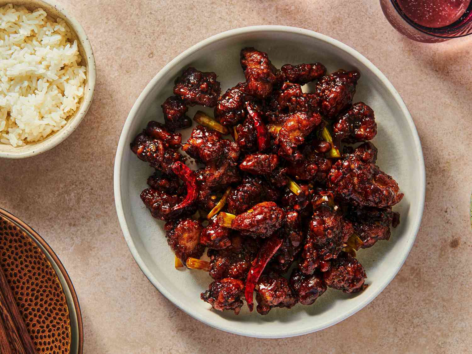 A wide ceramic bowl holding General Tso's chicken. In the upper left corner of the image is a ceramic bowl of white rice, and in the top right corner is a glass holding a beverage. There is another bowl in the bottom left corner.