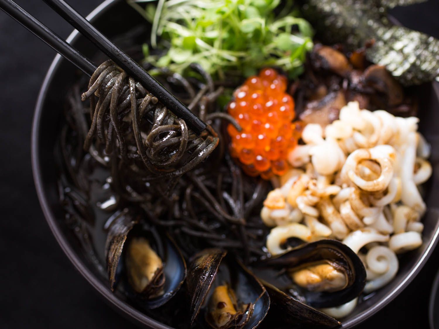 Chopsticks lifting a twirl of squid-ink spaghetti from a black bowl with mussels, squid, orange roe, micro greens, and nori