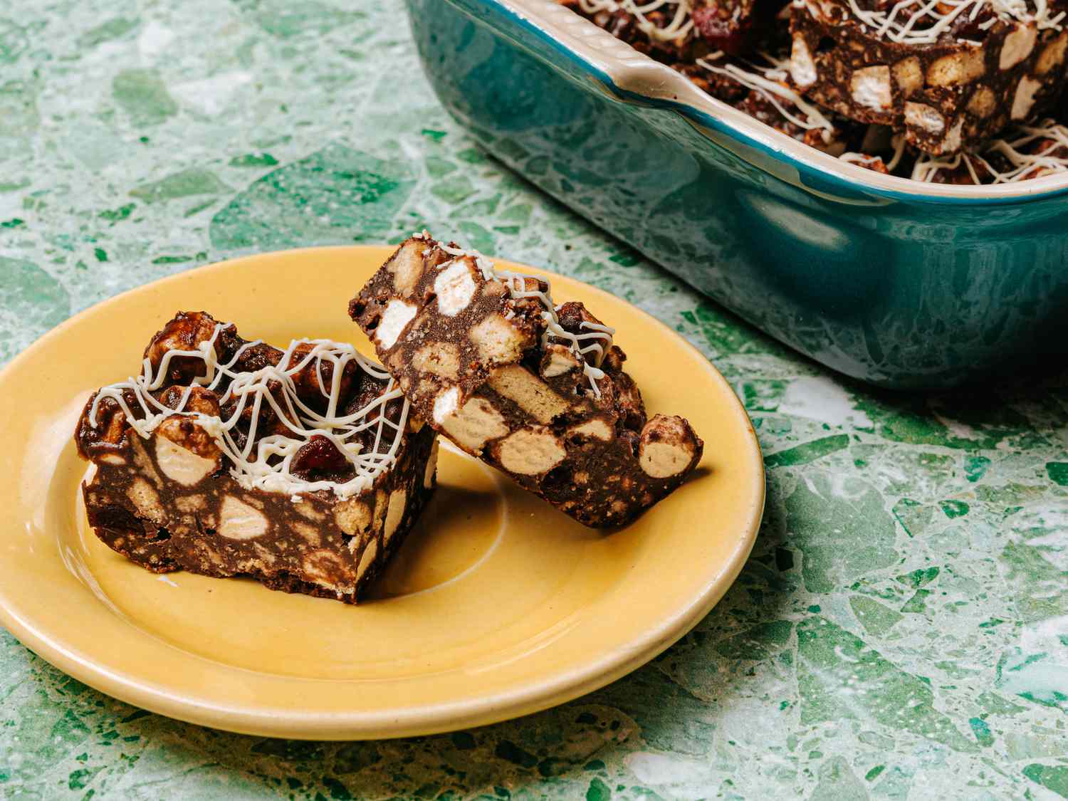 Rock Road squares on a yellow plate, and a green tarrazzo table, with a baking dish full of more rocky road